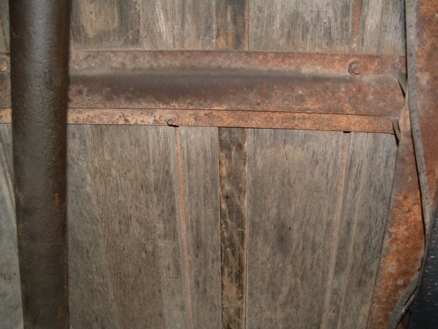 Underside wood detail.
Seen in the photo is the underside of one of the inverted hat channel crossmembers supporting the bed wood. Note the staggered bolt pattern through this channel. Where these bolts pass through the channel are located the wide joints between boards. These are the wide joints that are covered by the bed skid strips above. These wide joints can barely be seen in the photo. Also visible are other tighter joints. These are the gaps between the random width boards as they lay side by side. Note the difference in wood grain patterns and the how old black paint was absorbed and held differently by the various boards.
